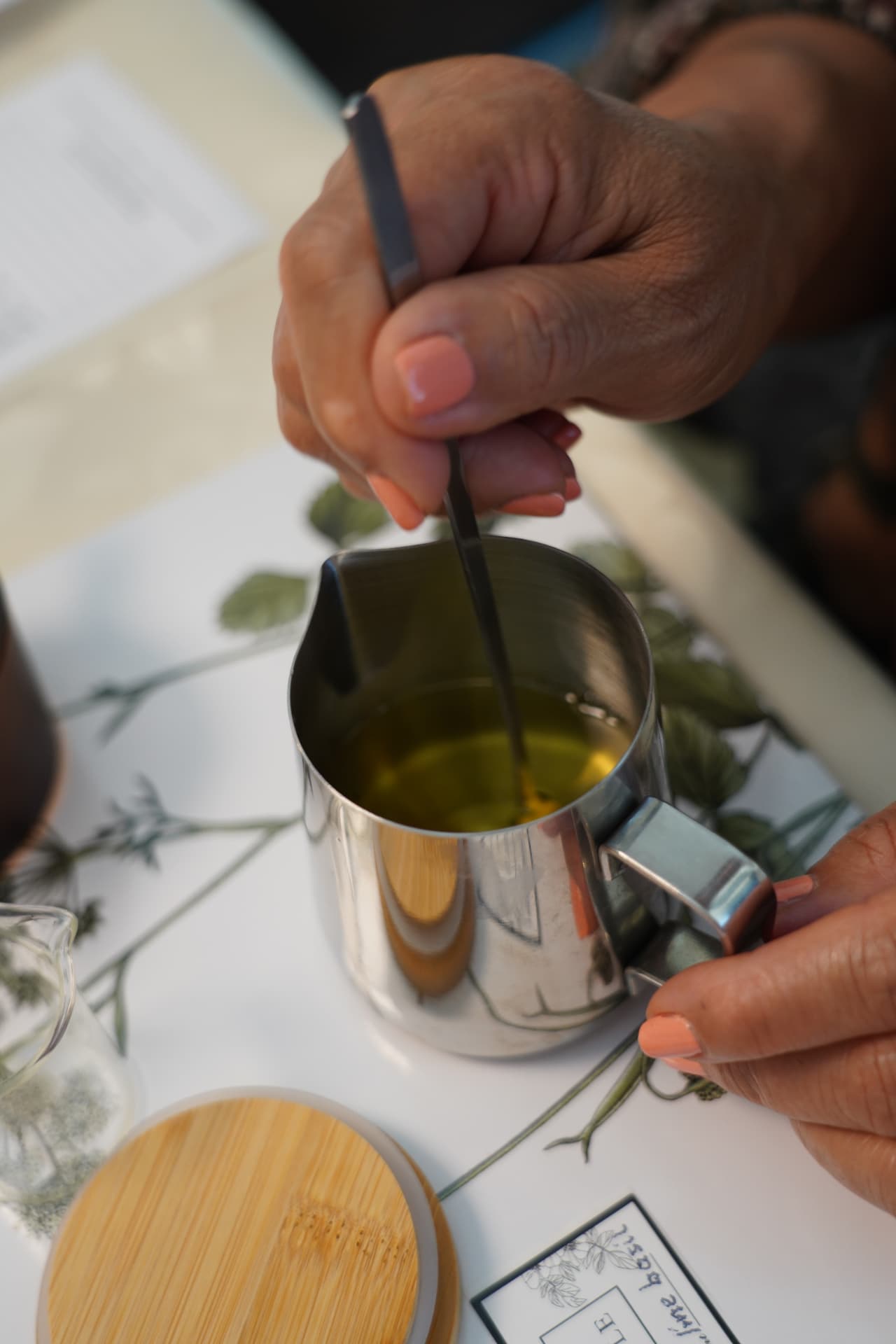 Hands stirring scented soy wax in a steel pouring jug over botanical prints