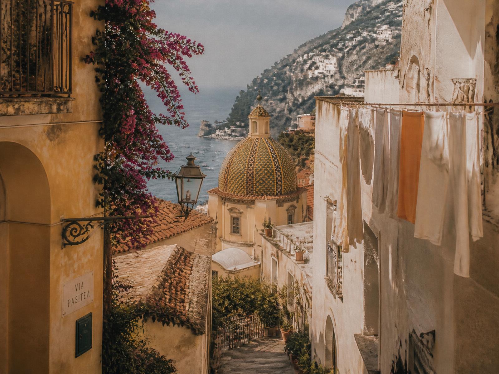 A pastel Amalfi street with a tiled dome and the sea beyond